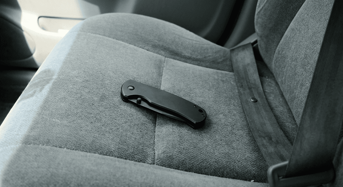 Navaja plegable negra cerrada sobre un asiento de tela gris de auto junto a un cinturon de seguridad